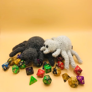 May include: Two crocheted spiders, one dark gray and one light gray, are surrounded by a collection of colorful polyhedral dice. The dice are in various colors, including green, red, yellow, and blue, and are scattered around the spiders.