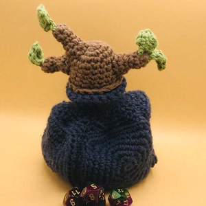 以下が含まれることがあります： 茶色の頭と緑色のアクセントが付いた手作りのかぎ針編みのフィギュアで、濃い青色のマントを着ています。紫と緑の色をした多面体のサイコロがいくつかフィギュアの前にあります。フィギュアは明るいオレンジ色の背景にあります。