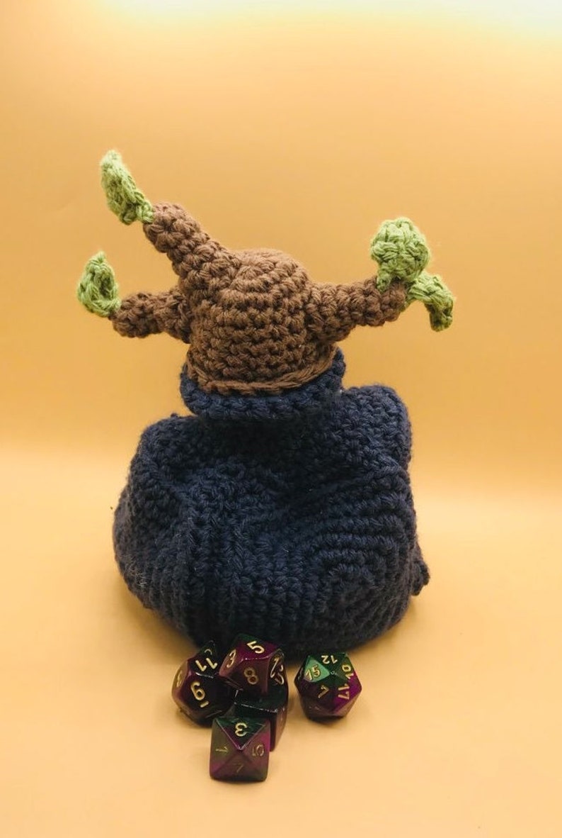 以下が含まれることがあります： 茶色の頭と緑色のアクセントが付いた手作りのかぎ針編みのフィギュアで、濃い青色のマントを着ています。紫と緑の色をした多面体のサイコロがいくつかフィギュアの前にあります。フィギュアは明るいオレンジ色の背景にあります。