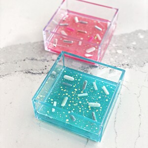 May include: Two square clear acrylic trays filled with colourful glitter and white sprinkles. The tray on the left is pink and the tray on the right is blue.