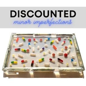 May include: A clear rectangular tray with a white background and a border of rolled up dollar bills. The tray is filled with colorful pills and capsules.