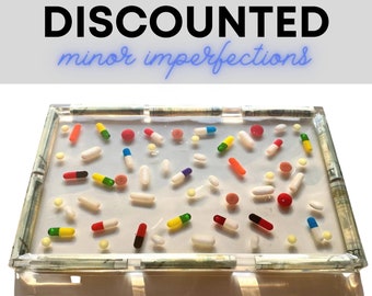 May include: A clear rectangular tray with a white background and a border of rolled up dollar bills. The tray is filled with colorful pills and capsules.