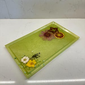 May include: A rectangular green tray with a light green border and a clear resin finish. The tray has dried flowers and a dried orange slice embedded in the resin.