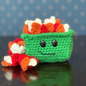 May include: A crocheted sushi roll set. The main piece is a green container with a smiling face and filled with orange, white, and red sushi. A separate sushi roll is next to the container. The background is a blue surface with white star shapes.