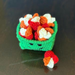 May include: A green crocheted container with a smiling face, filled with red, orange, and white crocheted food items. A small cluster of the food items sits outside the container. The container is handmade.