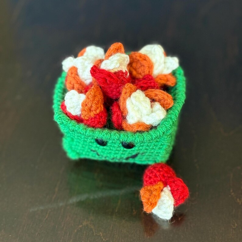 May include: A green crocheted container with a smiling face, filled with red, orange, and white crocheted food items. A small cluster of the food items sits outside the container. The container is handmade.