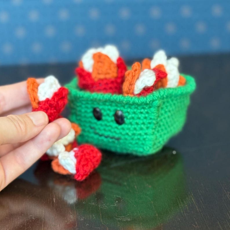 May include: A knitted sushi set, comprising a green container with a smiling face and several sushi pieces. The sushi is red, orange, and white. The set is displayed on a dark surface with a blue dotted background.