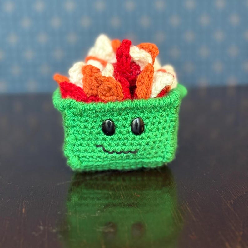 May include: A crocheted sushi roll in a green container with a smiling face. The sushi features white, orange, and red yarn. The container has button eyes and a stitched mouth. The background is a blurred blue pattern.
