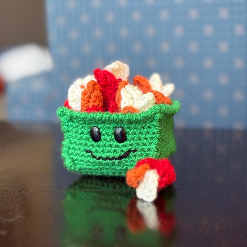 May include: A handmade, green crocheted sushi roll with a smiling face, filled with red, orange, and white yarn. A separate piece of sushi sits below. The background is a blurred blue with white dots.