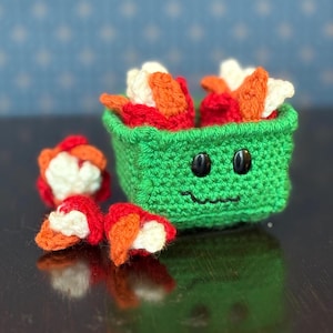 May include: A crocheted green container with a smiling face, filled with colorful sushi pieces. The sushi pieces are red, orange, and white. Several sushi pieces are scattered around the container. The background is a blue surface with white dots.