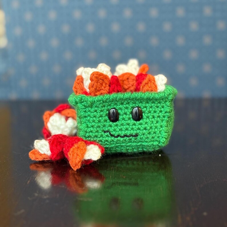 May include: A crocheted sushi roll set. The main piece is a green container with a smiling face and filled with orange, white, and red sushi. A separate sushi roll is next to the container. The background is a blue surface with white star shapes.