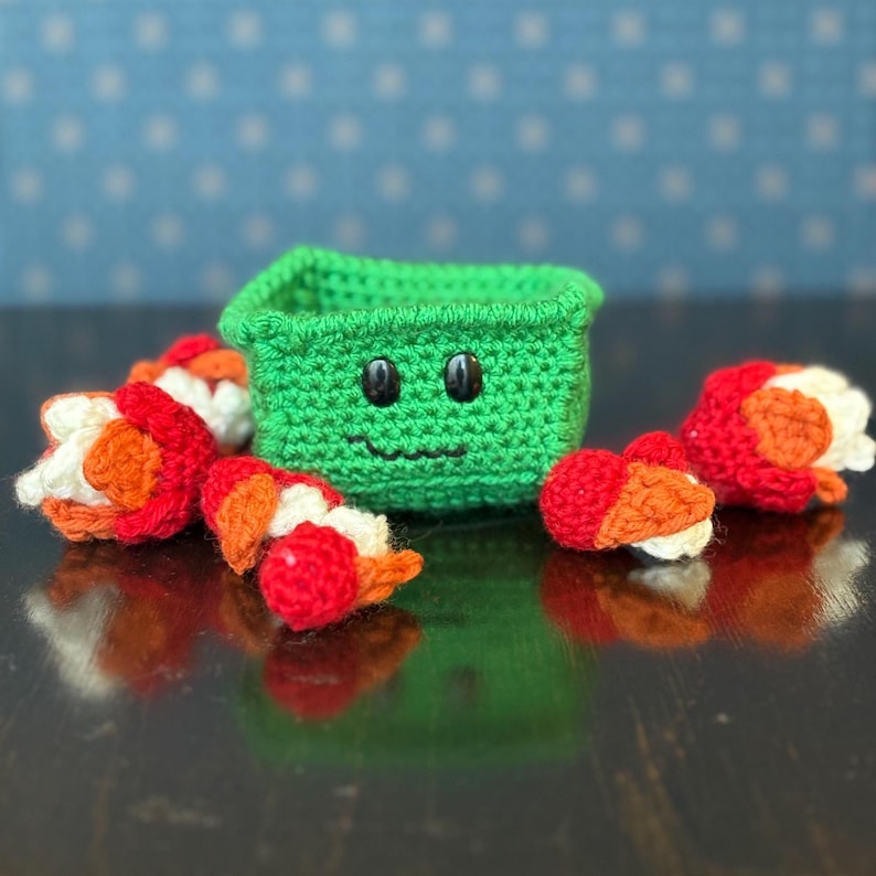 May include: A green crocheted box with a smiling face made of black buttons and yarn. Several crocheted sushi pieces with red, orange, and white details surround the box. The items are arranged on a dark, reflective surface.