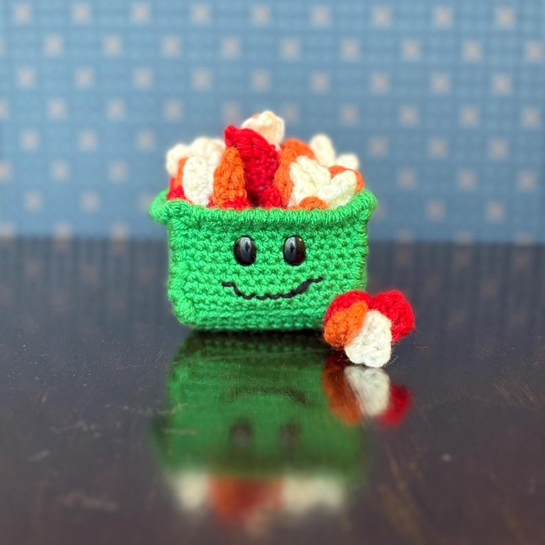 May include: A crocheted sushi set. A green container with a smiling face is filled with red, orange, and white sushi pieces. A small piece of sushi sits beside the container. The background is a blue dotted pattern.