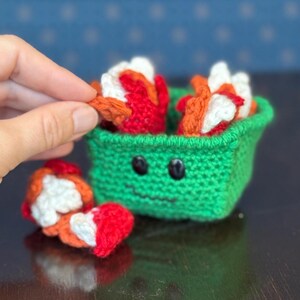 May include: A green crocheted container with a smiling face, holding several crocheted pieces in red, white, and orange. One piece is being held by a hand. The container is on a dark surface.