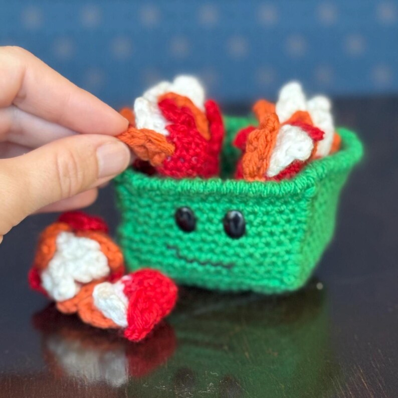 May include: A green crocheted container with a smiling face, holding several crocheted pieces in red, white, and orange. One piece is being held by a hand. The container is on a dark surface.