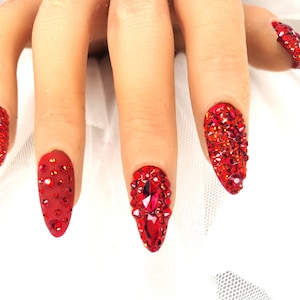 May include: Close-up of a hand with long, red, bejeweled artificial nails. Each nail is covered in small, sparkling red rhinestones, with larger gems on some. The nails are a vibrant, eye-catching accessory.