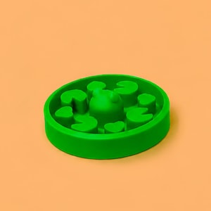 Può includere: Una ciotola circolare in silicone verde per l'alimentazione lenta degli animali domestici, caratterizzata da un motivo interno in rilievo con piccole forme arrotondate simili a foglie e una cupola centrale. Pareti lisce e base piatta.
