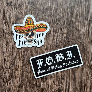 May include: Two stickers on a wooden surface. One sticker features a skull wearing a sombrero with the text "Fes Fier Sta". The other sticker is a black rectangle with the text "F.O.B.I. Fear of Being Included".