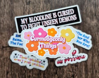 May include: A collection of colorful stickers with various phrases. One sticker reads "MY BLOODLINE IS CURSED TO FIGHT UNSEEN DEMONS." Another says "Curmudgeonly Things" with flower graphics. Other stickers have text like "Their opinions aren't your problem."