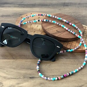 Puede incluir: Una cadena de gafas de cuentas de colores con cuentas turquesa, rosa, blancas y negras. La cadena está enroscada alrededor de unas gafas de sol negras.