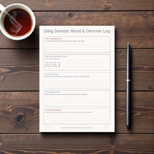 May include: A white Daily Somatic Reset & Glimmer Log journal with a black pen and a cup of tea on a wooden surface. The journal includes sections for morning baseline, nervous system state, daily glimmer, tool reflection, and evening release.