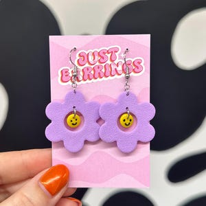 May include: Purple flower-shaped earrings with a yellow smiley face charm. The earrings are on a pink card with the text "JUST EARRINGS".