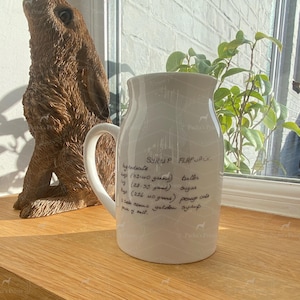May include: A white ceramic pitcher featuring black handwritten text that reads Syrup Flapjack, listing ingredients including 4 oz (113.40 grams) butter, 1 oz (28.35 grams) sugar, 8 oz (226.40 grams) porridge oats, 2 tablespoons golden syrup, and a pinch of salt.