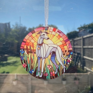 May include: A circular hanging ornament featuring a stained-glass style illustration of a white greyhound standing in a field of flowers and tall grass. The background depicts a vibrant orange and yellow sunburst. It hangs from a white ribbon.