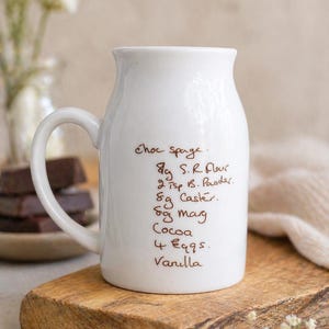 May include: A white ceramic jug with a handle, inscribed with a handwritten chocolate sponge recipe. Ingredients include self-raising flour, baking powder, caster sugar, margarine, cocoa, eggs, and vanilla. The jug rests on a wooden board.