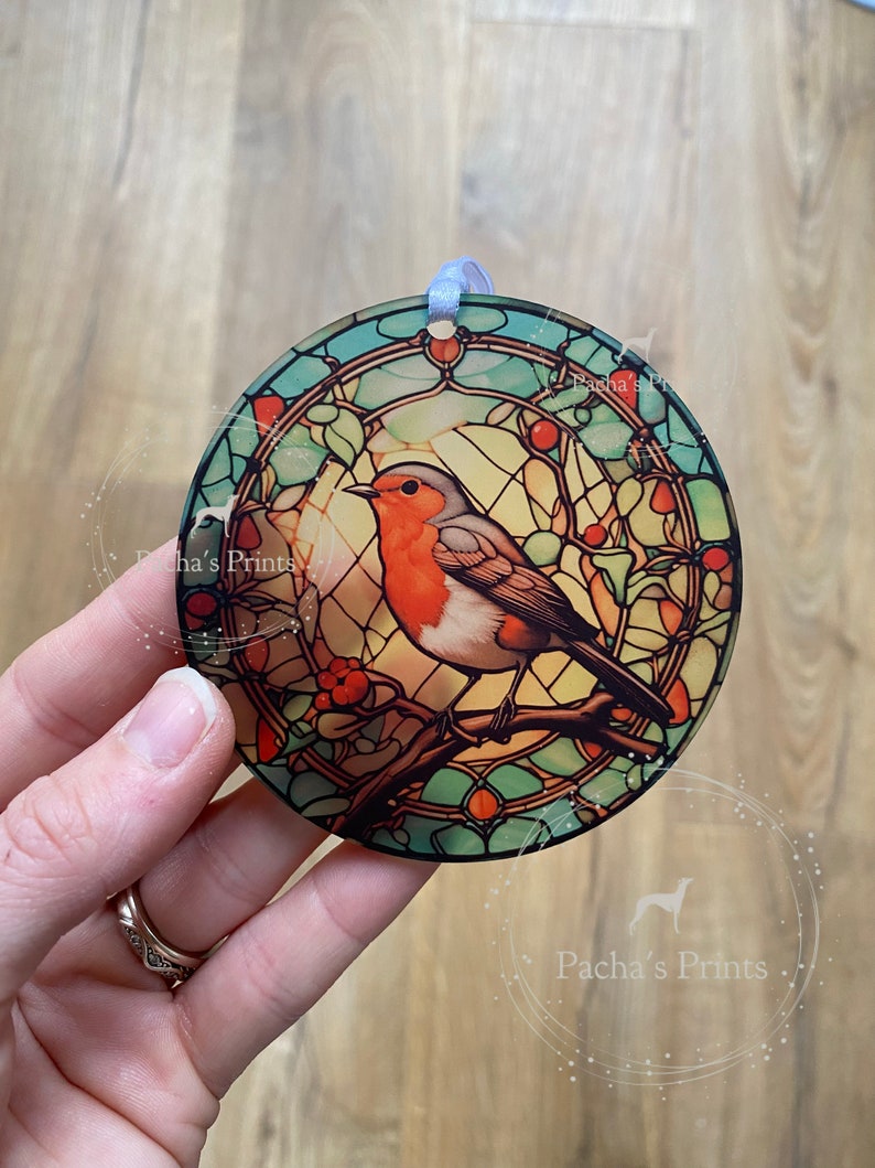May include: A round, stained glass ornament featuring a robin perched on a branch with red berries. The ornament has a blue ribbon attached to the top.