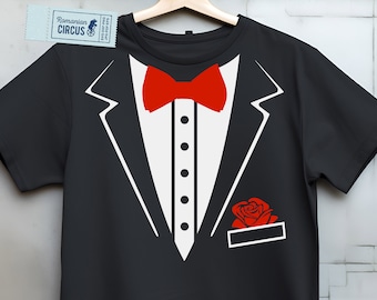 May include: Black t-shirt with a tuxedo design. The design includes a white shirt front, a red bow tie, and a red rose in the pocket. The shirt is hanging on a wooden hanger.