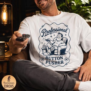 May include: A white t-shirt featuring a dark blue graphic of a person sitting in an armchair holding a remote control. The text reads Professional BUTTON PUSHER, with a small mug labeled DAD on a side table, surrounded by decorative flourishes.