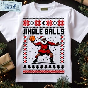 May include: White t-shirt with a Christmas-themed graphic. The design features a Santa Claus figure in a basketball pose, holding a basketball. The text "JINGLE BALLS" is above the Santa illustration. The shirt has a festive, patterned border.