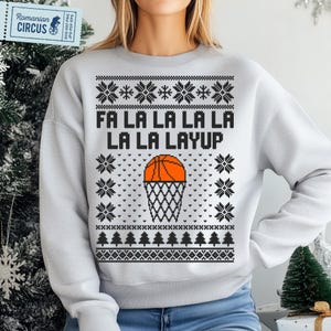 May include: A light gray sweatshirt with a basketball-themed Christmas design. The design includes the text "FA LA LA LA LA LA LA LAYUP", a basketball, and a net. The sweatshirt also features snowflake and tree patterns.