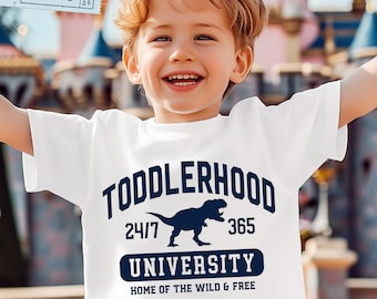 May include: A white short-sleeved t-shirt featuring dark blue text that reads TODDLERHOOD 24/7 365 UNIVERSITY HOME OF THE WILD & FREE, with a silhouette of a Tyrannosaurus Rex dinosaur positioned in the center of the graphic design.