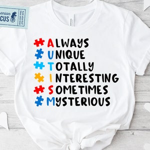 Puede incluir: Camiseta blanca con un gráfico negro que dice "#ALWAYS #UNIQUE #TOTALLY #INTERESTING #SOMETIMES #MYSTERIOUS" con piezas de rompecabezas de diferentes colores detrás de cada letra.