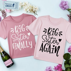 May include: Two pink t-shirts with the text "Big Sister" on them. One shirt says "Finally" and the other says "Again".