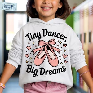 May include: White hoodie featuring the text "Tiny Dancer, Big Dreams" and a graphic of pink ballet shoes with a bow. The design includes hearts and stars. The hoodie is worn by a person.