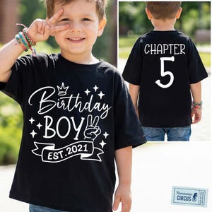 May include: Black t-shirt with white text reading "Birthday Boy EST. 2021" and "Chapter 5". The front of the shirt features a crown and peace sign graphic. The back of the shirt displays the number 5.