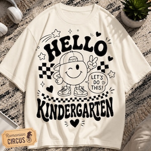 May include: A cream-coloured t-shirt featuring a black graphic print. The design includes a winking smiley face wearing a baseball cap, surrounded by stars, hearts, and checkerboard patterns. The text reads HELLO KINDERGARTEN and LET'S DO THIS!