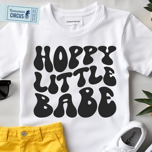 Może przedstawiać: Biała koszulka z czarnym napisem "Hoppy Little Babe" w retro, groovy czcionce.