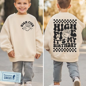 Op de afbeelding: Een crèmekleurige trui met zwarte tekst die "High Five It's My Birthday" zegt met een zwart-wit geruit patroon op de rug. De voorkant van de trui heeft een zwarte hand met de tekst "High Five" eromheen.