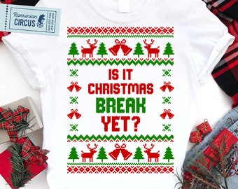 May include: White t-shirt with a festive Christmas design. The design includes red reindeer, green Christmas trees, and red bells. The text "IS IT CHRISTMAS BREAK YET?" is in red and green. The shirt has a classic crew neck.