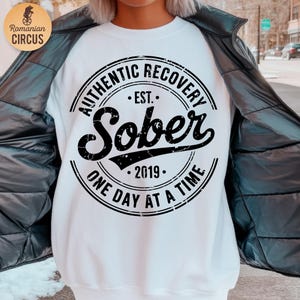 Könnte beinhalten: Weißes Sweatshirt mit einem schwarzen kreisförmigen Aufdruck, der "AUTHENTIC RECOVERY EST. SOBER 2019 ONE DAY AT A TIME" zeigt. Das Sweatshirt wird unter einer schwarzen Jacke getragen. Das Logo "Romanian CIRCUS" befindet sich oben links.