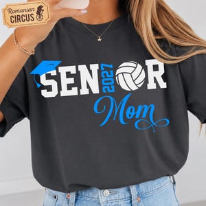 Pu&ograve; includere: Una t-shirt grigio antracite con testo grafico bianco e blu che recita SENIOR 2027 MOM. Il design include un'icona di tocco di laurea blu che sostituisce la lettera I e una grafica di pallavolo bianca che sostituisce la lettera O in SENIOR.