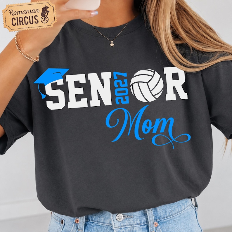 Pu&ograve; includere: Una t-shirt grigio antracite con testo grafico bianco e blu che recita SENIOR 2027 MOM. Il design include un'icona di tocco di laurea blu che sostituisce la lettera I e una grafica di pallavolo bianca che sostituisce la lettera O in SENIOR.