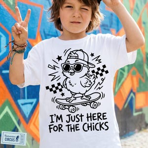 Könnte beinhalten: Weißes T-Shirt mit einer schwarzen Grafik eines Küken mit Sonnenbrille und Kappe auf einem Skateboard. Der Text "I'M JUST HERE FOR THE CHICKS" steht darunter. Der Hintergrund ist eine bunte Graffiti-Wand.