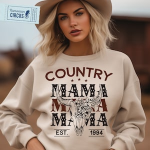 May include: A beige sweatshirt with a brown and black graphic design that says "COUNTRY MAMA MAMA EST. 1994". The graphic includes a skull with flowers and antlers.
