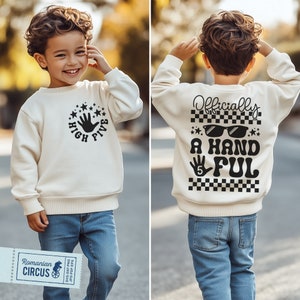 Op de afbeelding: Een wit sweatshirt met een zwarte afbeelding van een hand die een high five geeft met de tekst "High Five" eromheen. De achterkant van het sweatshirt heeft een zwarte afbeelding van een hand met de tekst "Officially A Handful" met een zonnebril en een geruit patroon.