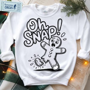 May include: White sweatshirt with a black and white graphic of a gingerbread man with a broken leg and the words "Oh Snap!". The design is a humorous take on a classic holiday theme.